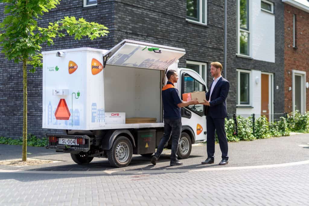 PostNL employee delivers a parcel to a man wearing a suit in front of an open PostNL truck in a residential area PostNL bezorger overhandigt pakket aan man